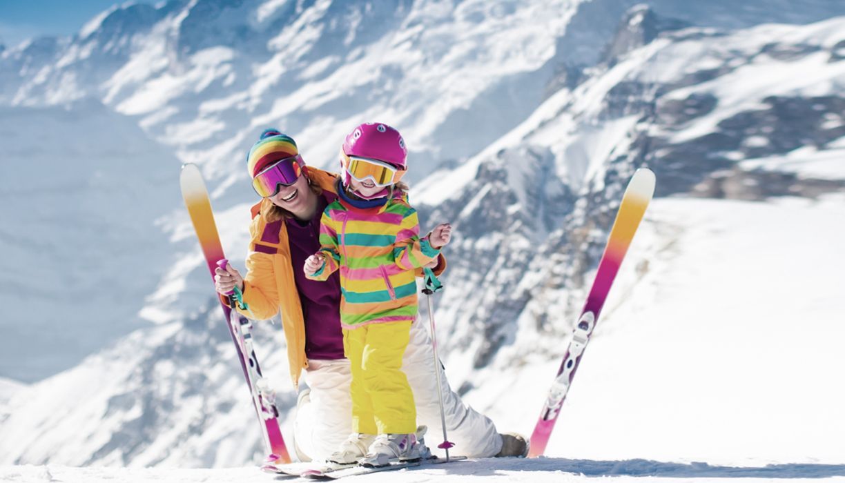 Mutter und Kind mit Ski und Skibrille lachen in die Kamera, dahinter die verschneite Bergflanke