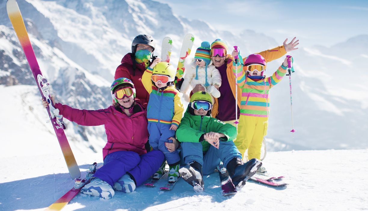 Familie mit vier Kindern sitzt lachend im Schnee, Ski und Stöcke in der Hand