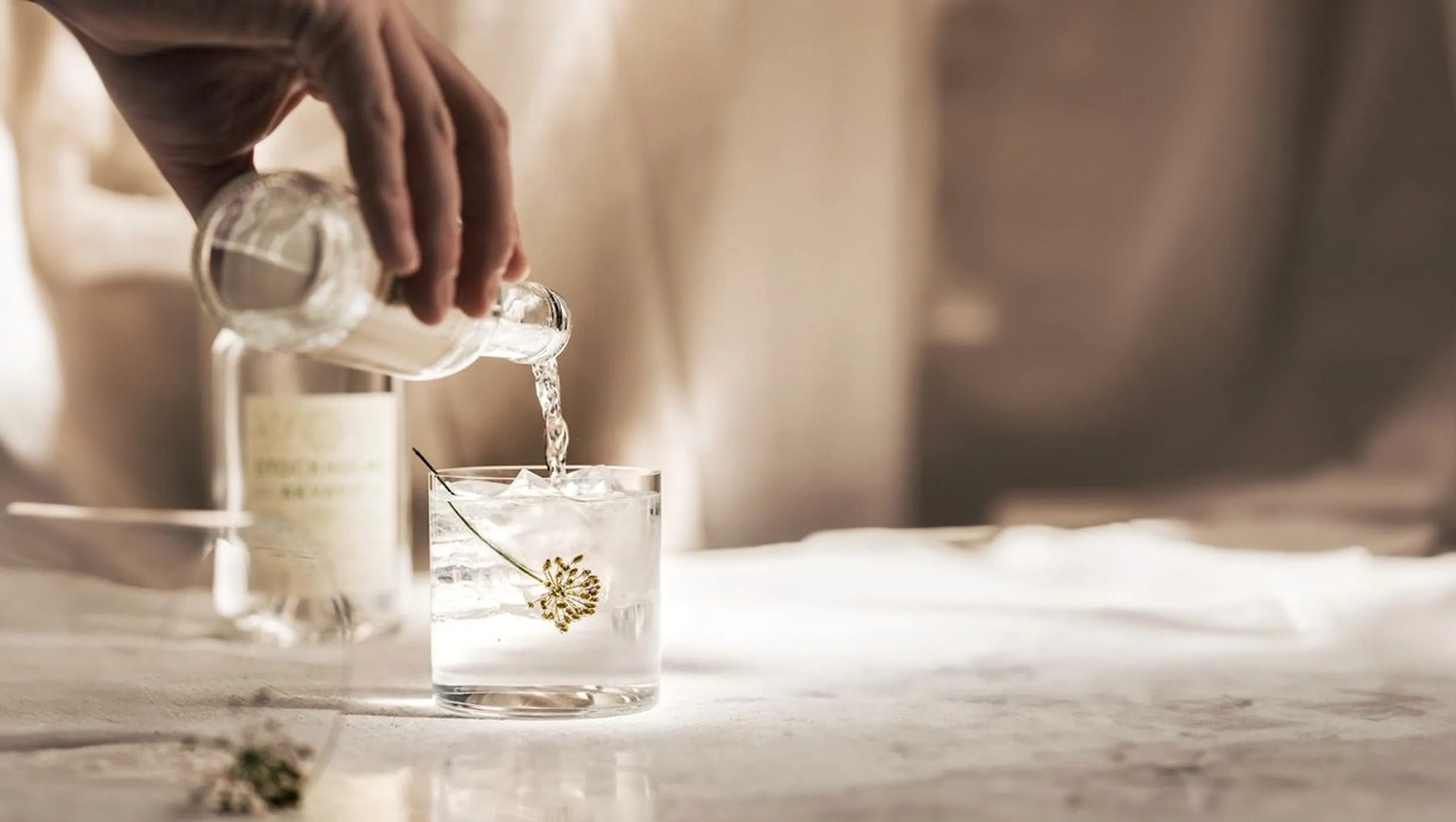 Eine Hand schenkt aus einer Flasche in ein Glas mit Eis ein, im Glas schwimmt eine Blüte