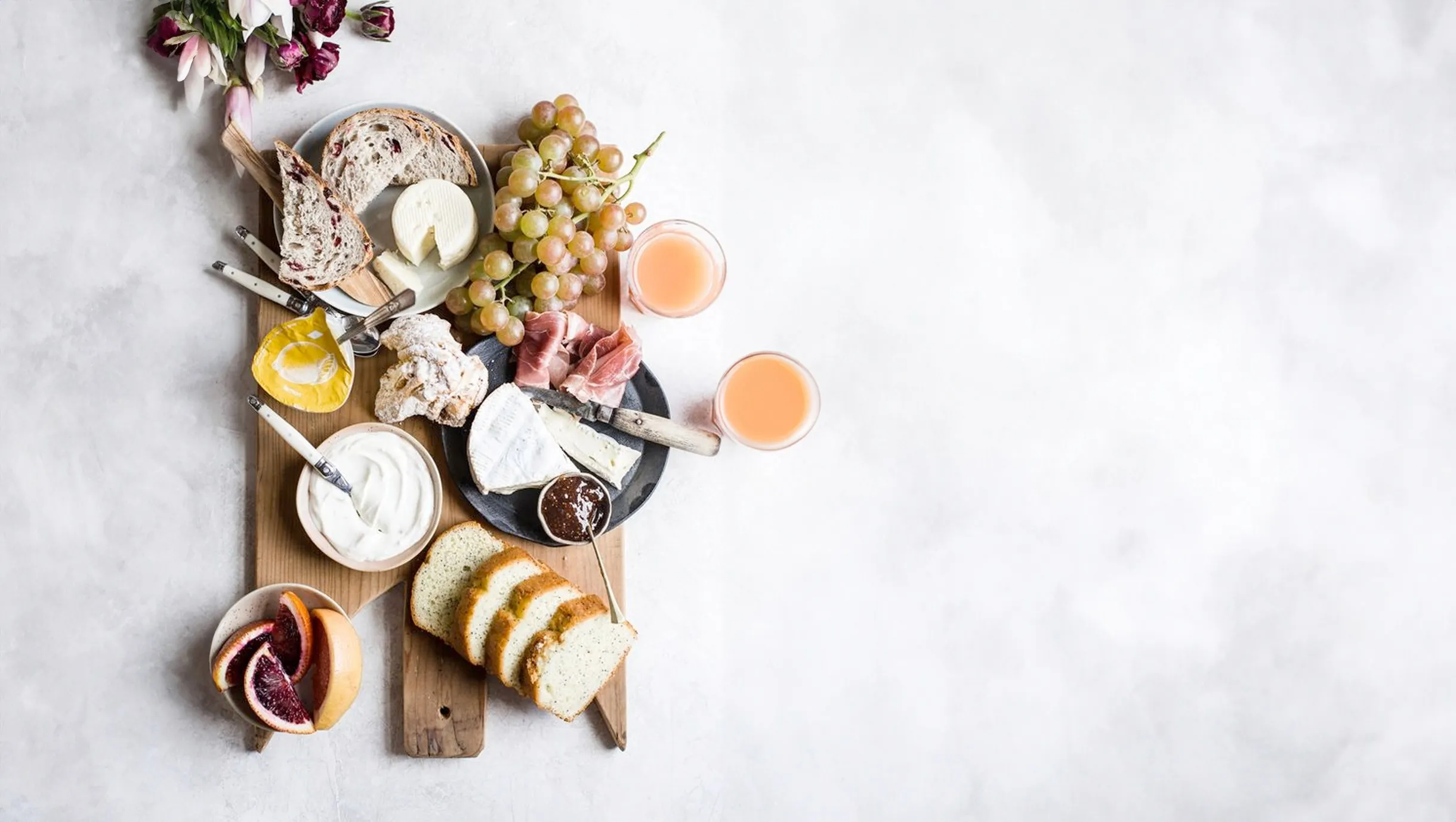 Frühstücksbrett mit Brot, Käse, Schinken, Weintrauben und zwei Gläsern Saft