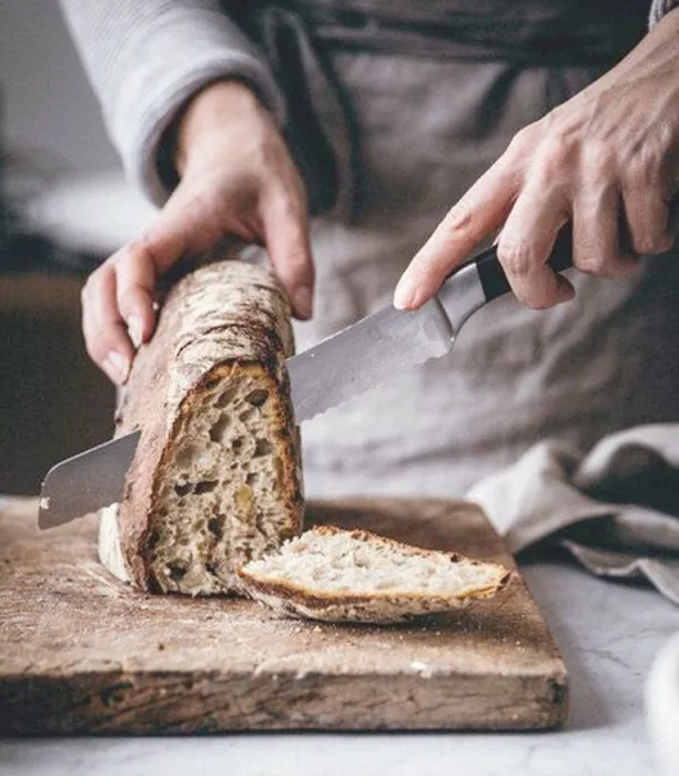 Hände schneiden ein rustikales Sauerteigbrot mit langem Messer auf einem Holzbrett