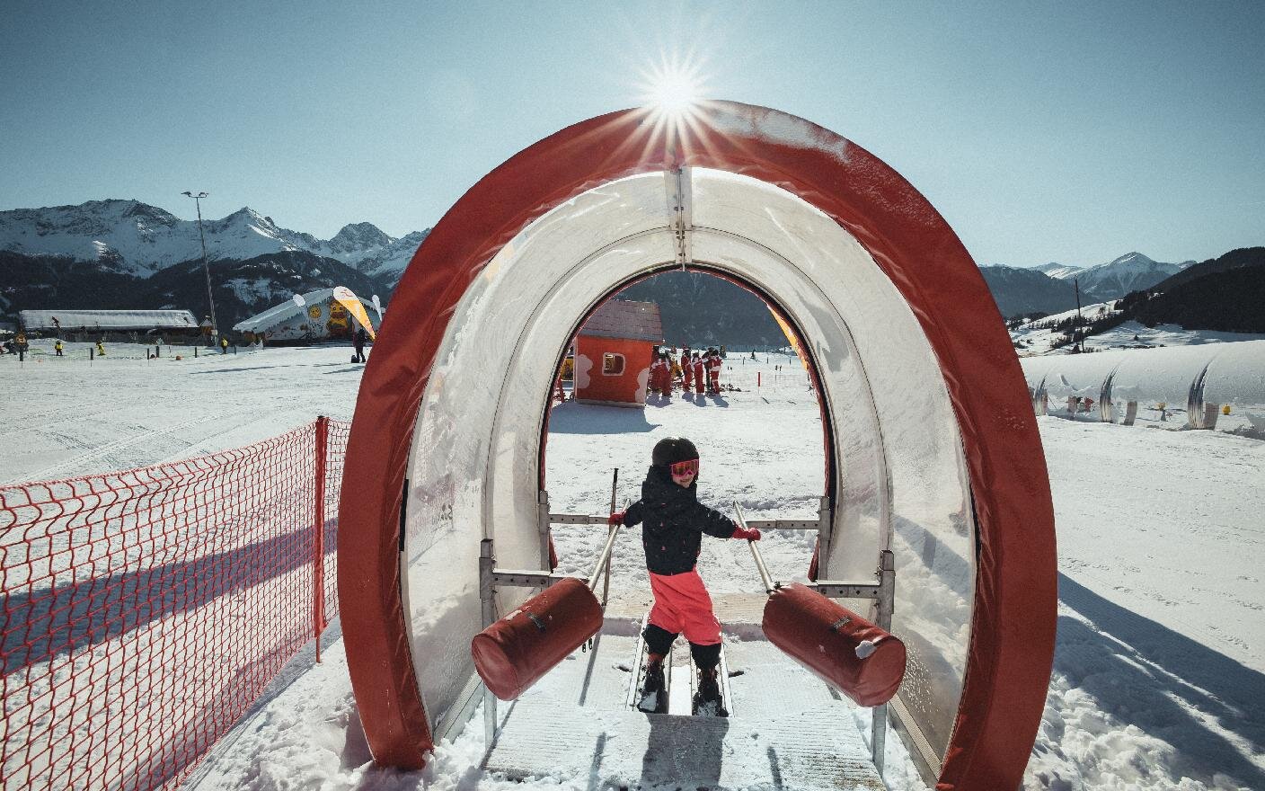 Kind auf Ski fährt durch den roten Tunnelbogen der Übungsanlage, dahinter die Bergkette