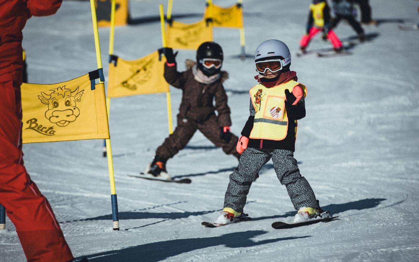 Zwei Kinder im Skikurs zwischen gelben Berta-Fahnen, eines mit der Warnweste der Skischule