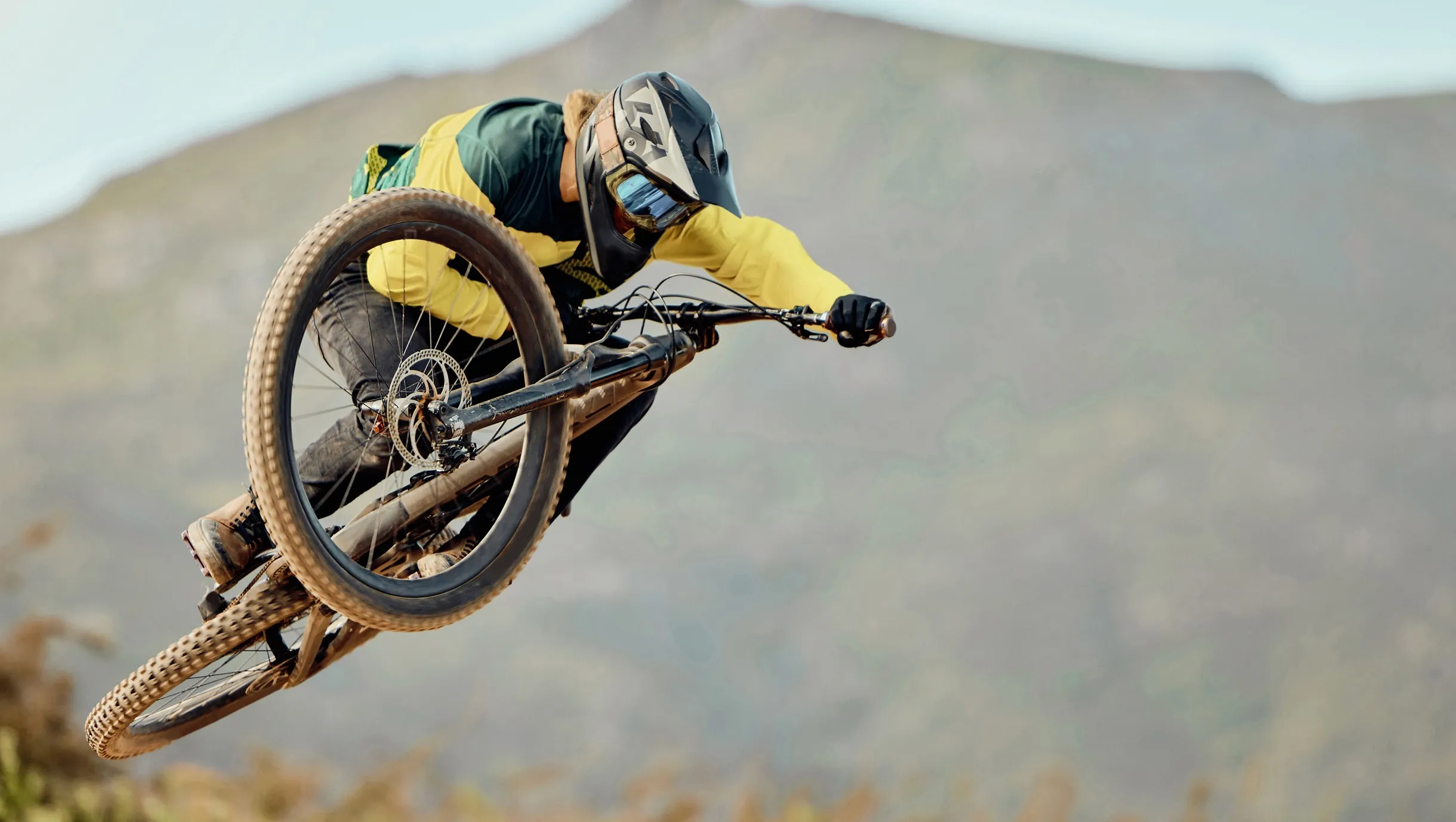 Mountainbiker in gelber Jacke springt mit dem Rad vor einer steilen Felswand.