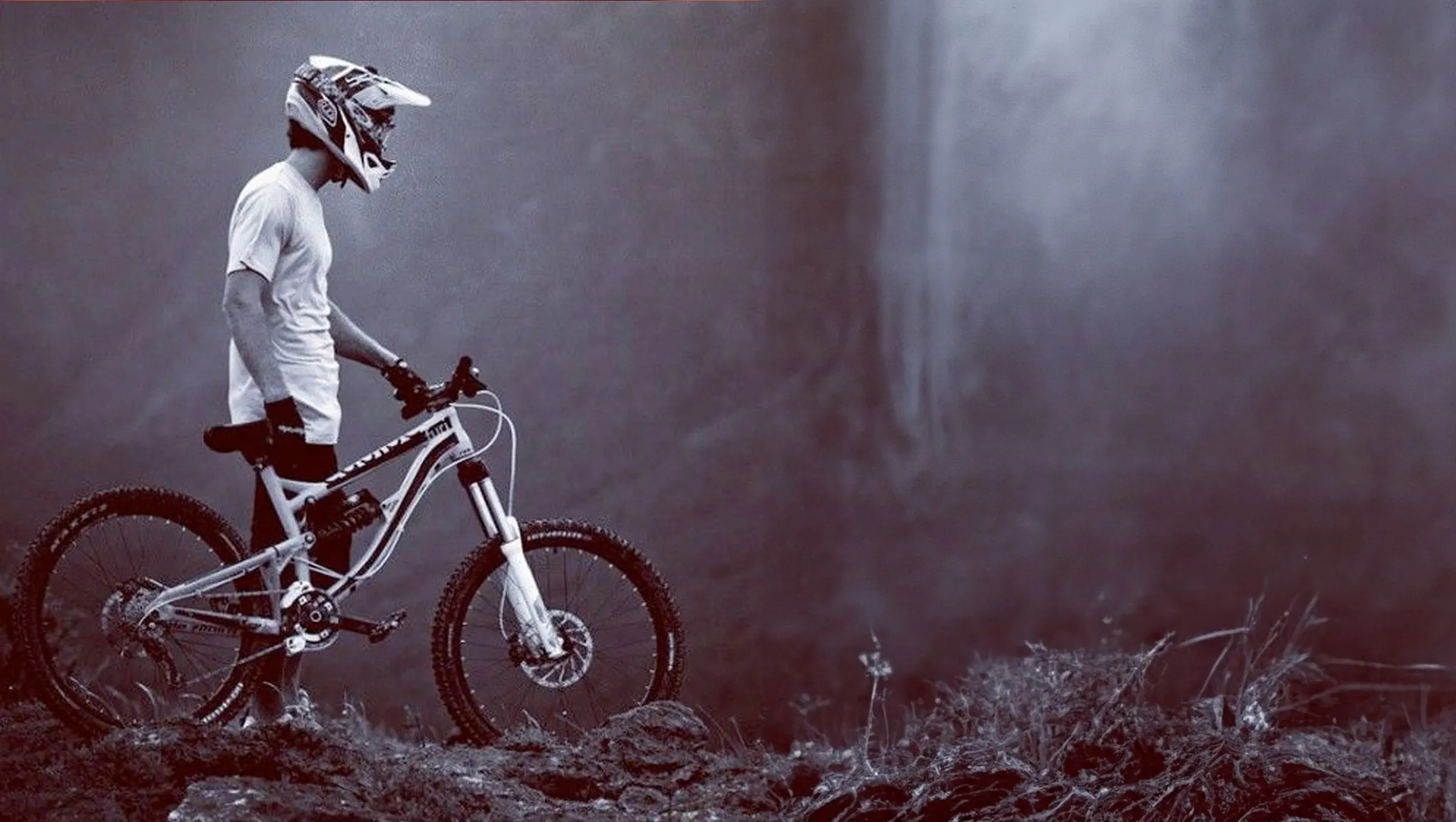 Fahrer mit Fullface Helm steht mit seinem Downhill Rad im Nebel vor dunklen Bäumen.