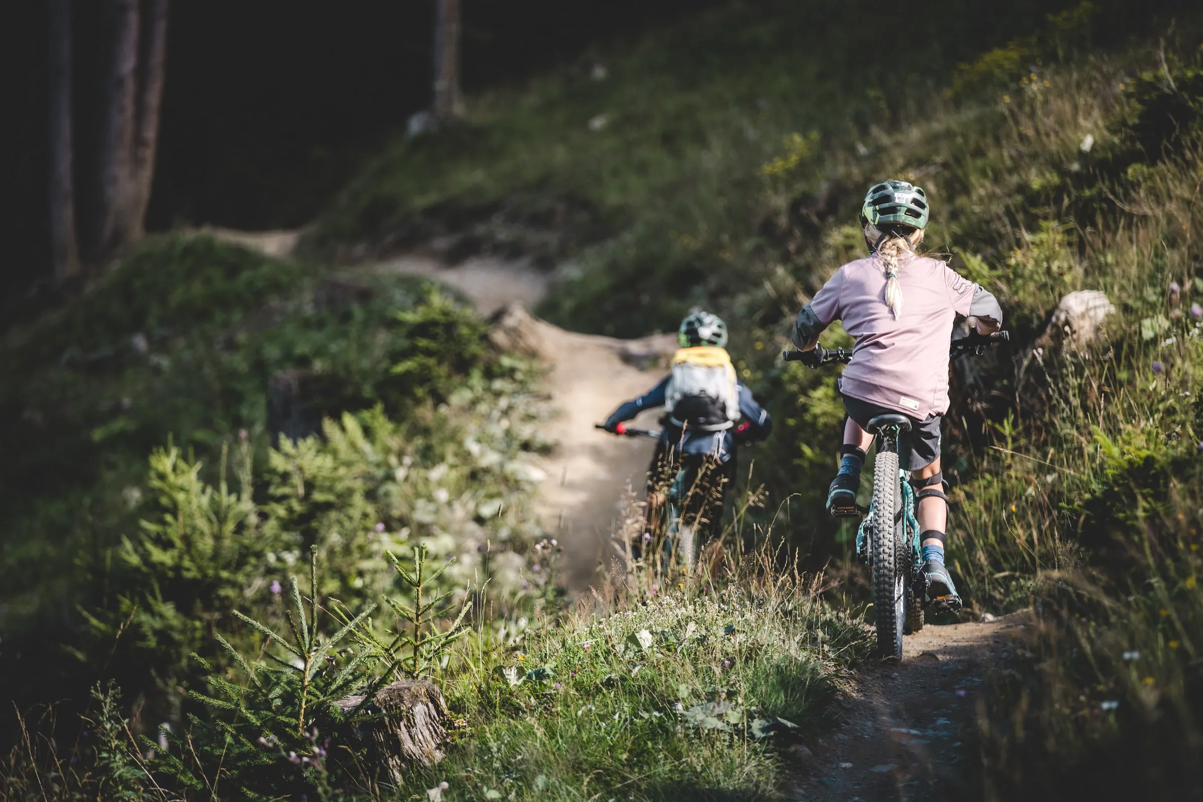 Zwei Mountainbiker fahren hintereinander über einen grünen Trail, vorne fährt ein Kind.