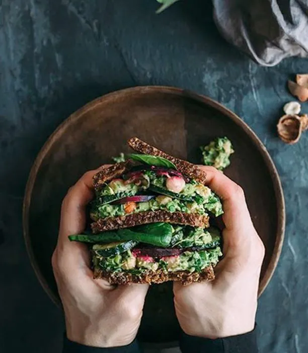 Zwei Hände halten ein belegtes Vollkornbrot über einer runden Holzschale.
