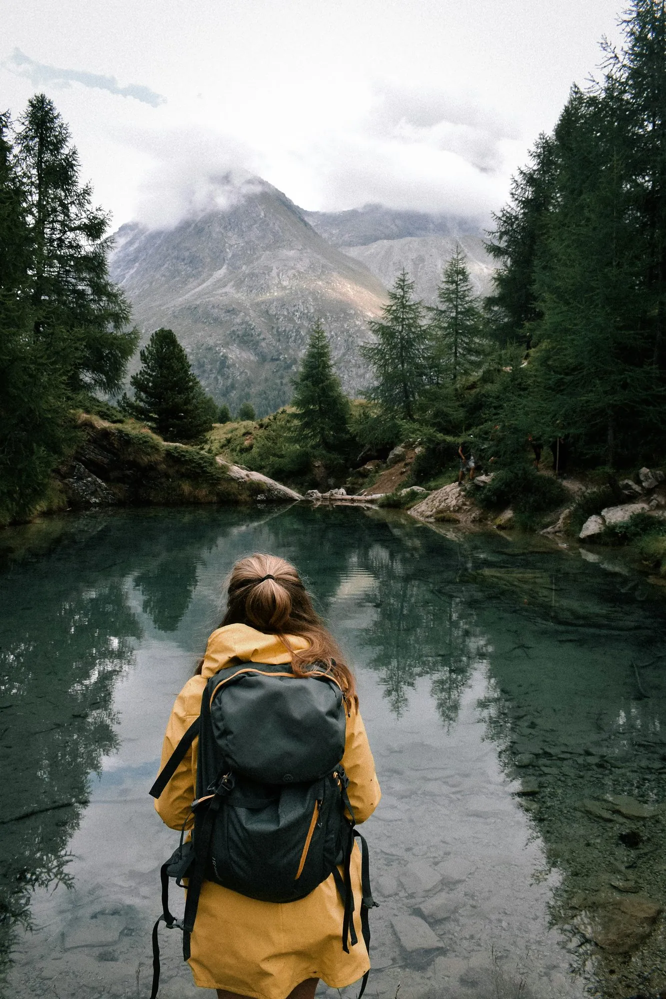 Wanderin mit gelber Jacke am Ufer eines Bergsees im Lärchenwald