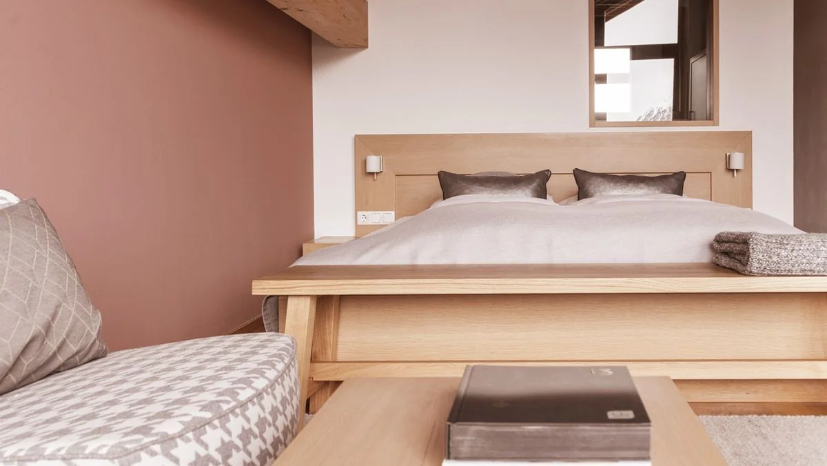 Bett mit Holzkopfteil vor altrosa Wand, Bank davor, rechts der offene Badbereich